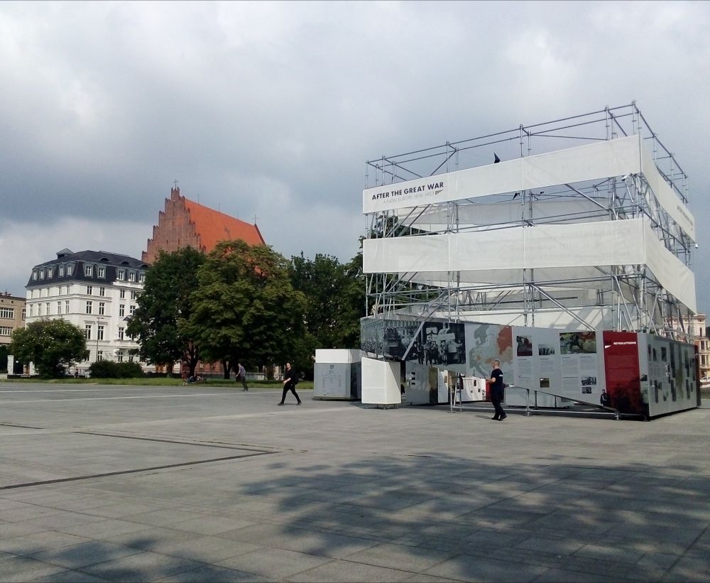 Cloudy day. The cube-like installation of the After the Great War exhibition stands in the middle of an empty square. On its left, in the background greenery and gothic red-brick church.