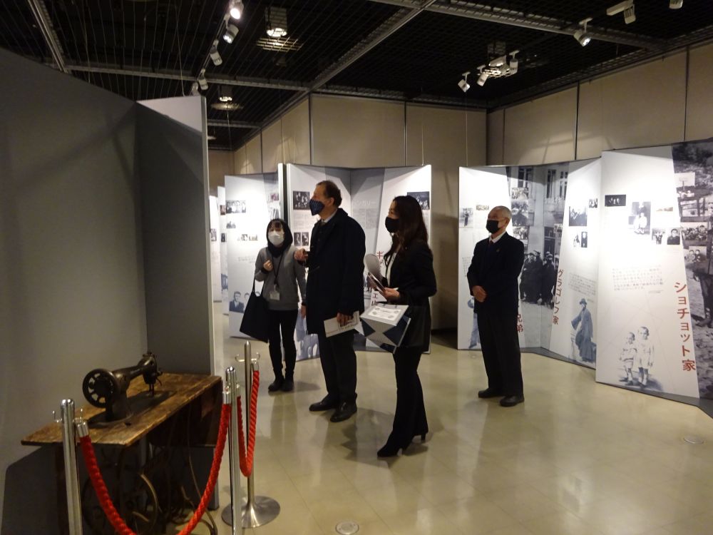 Italian Consul General Luigi Diodati, accompanied by two women and one man, looks attentively at one of the panels of the Between Life and Death Exhibition. surrounding him, other panels with black and white photographs. On his left, a vintage sawing machine is exhibited.