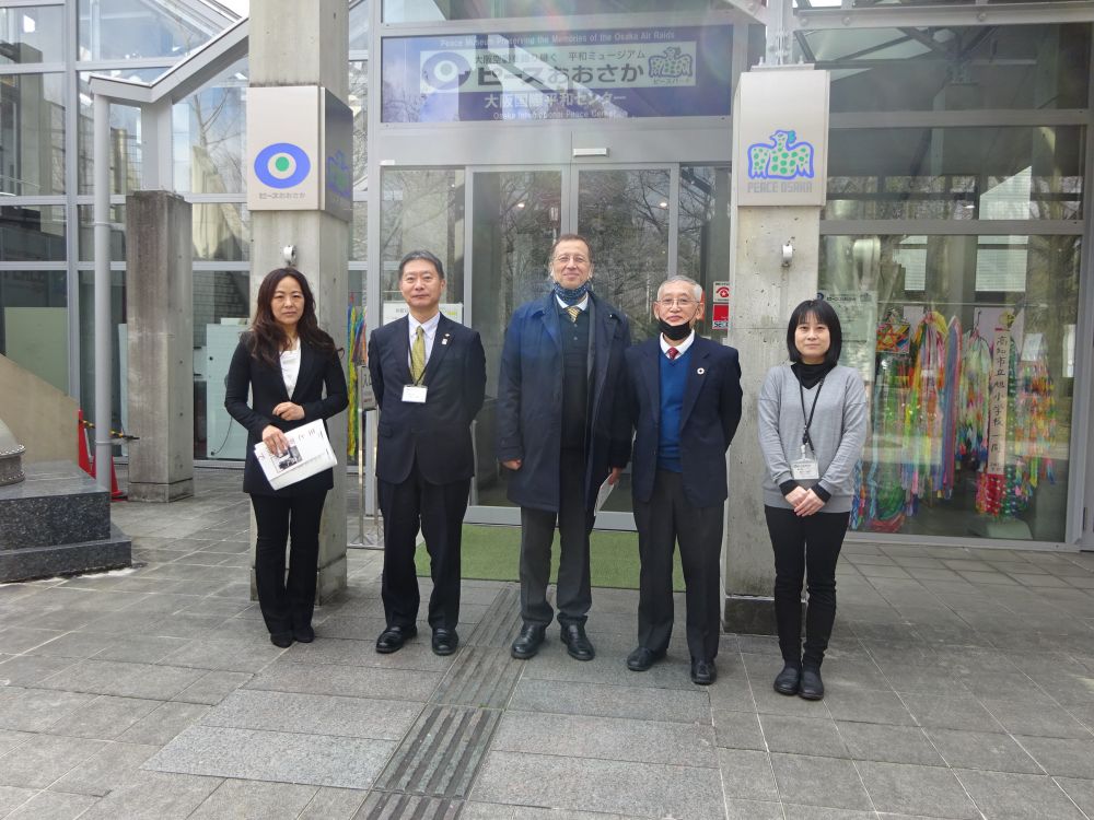 Cloudy day. A group of five well-dressed dignitaries, two women and three men, stand in front of a building, smiling. The banner on the glass-and-steel facade of the building reads Osaka International Peace Centre