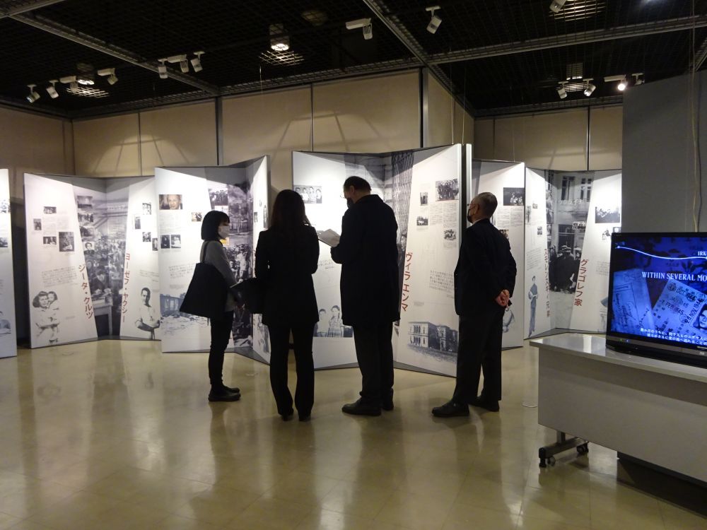 Interior. Italian Consul General Luigi Diodati, accompanied by two women and one man, stands in front of panels of the Between Life and Death Exhibition. surrounding him, other panels with black and white photographs. On his right, a TV set plays a video connected to the themes of the exhibition.
