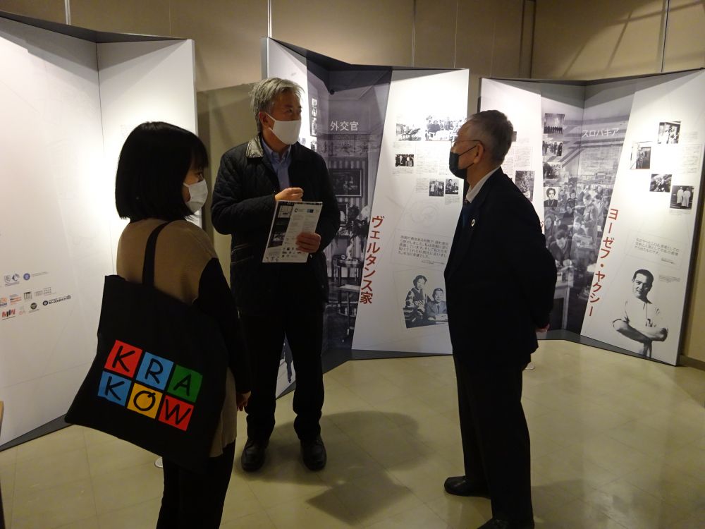 Interior. Richard Mei, Consul General of the US, accompanied by a woman and man, stands in front of panels of the Between Life and Death Exhibition. He is engaged in a conversation with his companions.