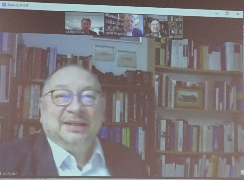 Pull down projector screen. Displayed on it, Profesor Jan Rydel sits in front of a wall of bookshelves. He is videoconferencing through Zoom with several other speakers.