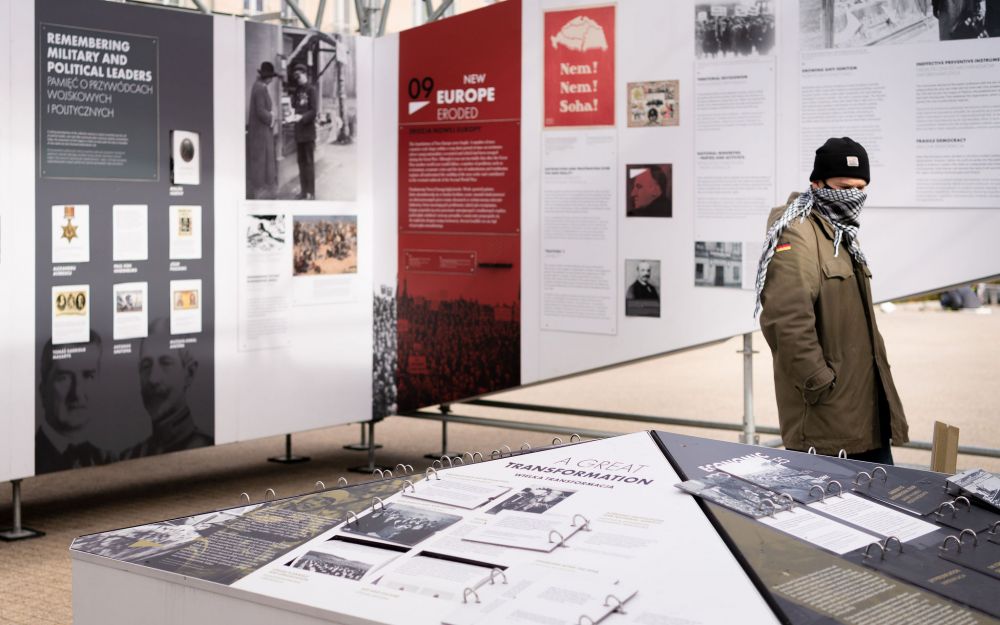 Interior of the installation of After the Great War exhibition. In the center, a knee-high board with the infographics, surrounded from all sites by the walls of the exhibition. A single young man looks at  something on a board.