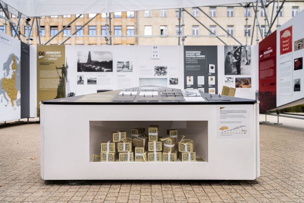 Interior of the installation of After the Great War exhibition. In the center, a knee-high board with the infographics, surrounded from all sites by the walls of the exhibition. Beneath the board, on the level of visitors knees, a display case shows stacks of banknotes, illustrating the post-war hyperinflation.