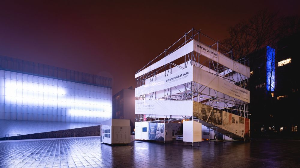 Night. The empty towns square is illuminated by the strong neons on the facade of the building on the left. On the right stands the cube-like installation of the After the Great War exhibition.