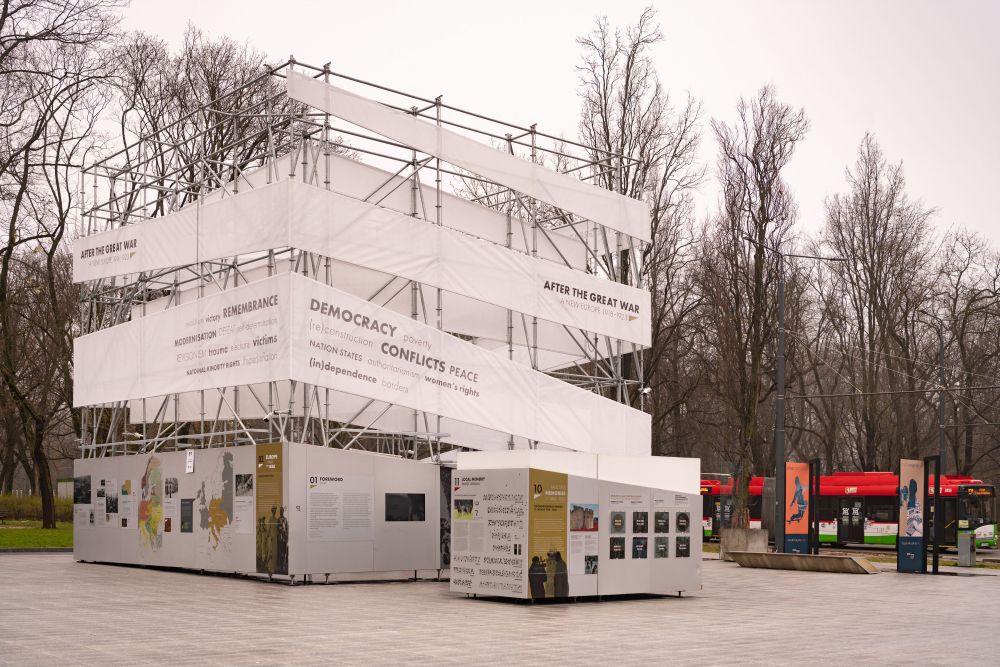 Cloudy day. The cube-like installation of the After the Great War exhibition stands in the empty square. In the background, a row of leafless trees.
