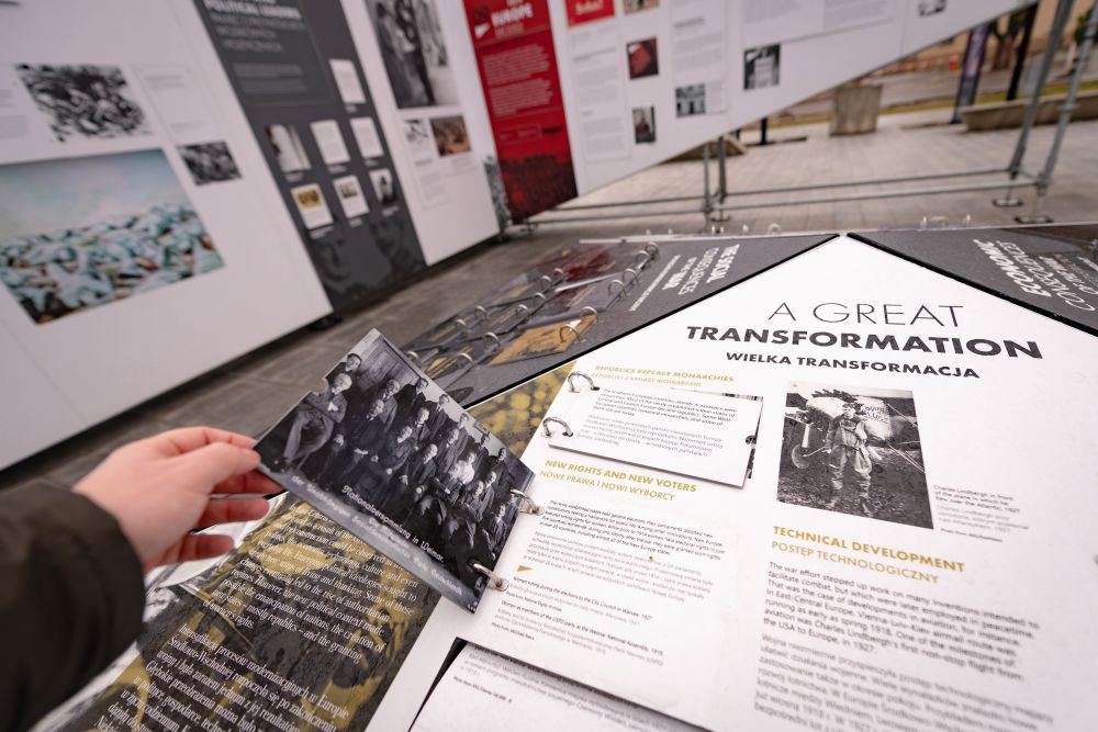 A disembodied hand flips through the flip-deck bind to the central board of the After the Great War exhibition. The panel in front of them contains information on the social transformations and technological progress during the interwar period. Fliped flip-deck reveals an old black-and-white photography of well-dressed older men.