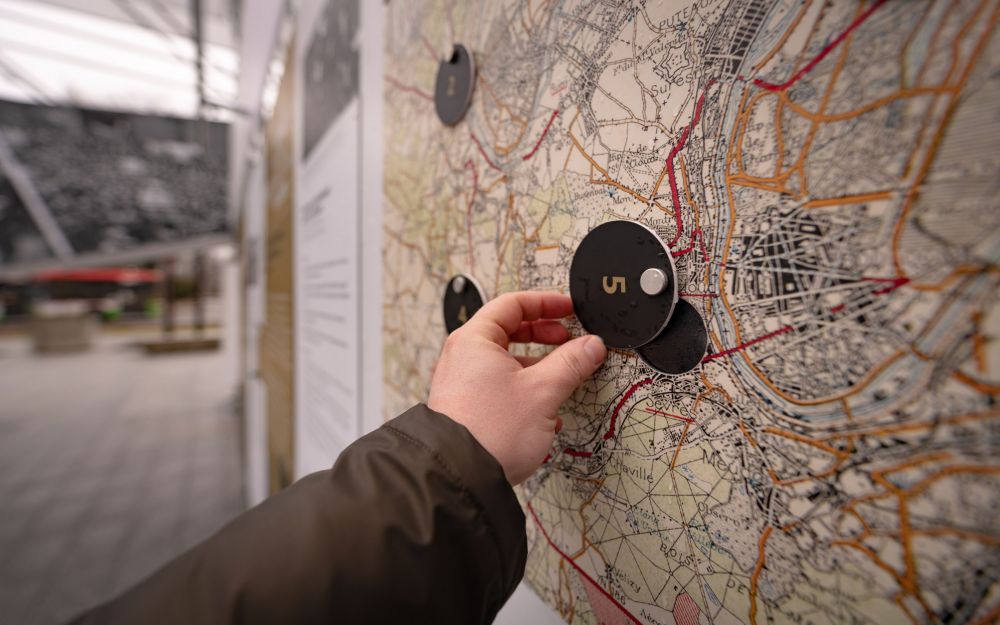 A disembodied hand lifts the peephole placed in the wall of the exhibition.   The peephole is placed on a wall that presents the map of Paris and its surroundings during the time the negotiations of the treaty of Versailles took place.