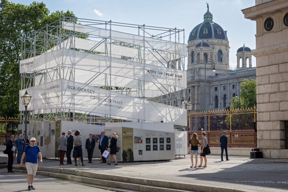 Sunny day.  Cube-like installation of the After the Great War exhibition stands in the square, groups of people gather around it. Behind it, decorative black and white fence. Further in the background, a dome of a neoclassical palace.