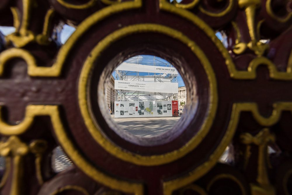 A black-and-gold fence surrounding Heldensplatz. Through the eye of one of the rings of the fence one can see the cube-like installation of the After the Great War exhibition.