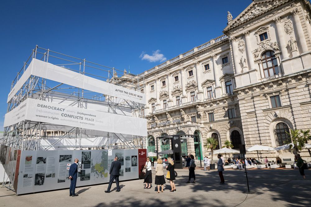 Sunny day. On the left, cube-like installation of the After the Great War exhibition stands in the square, groups of people gather around it, reading from infographics presented on its walls. On the right, the neo-classical architecture of Hofburg palace.
