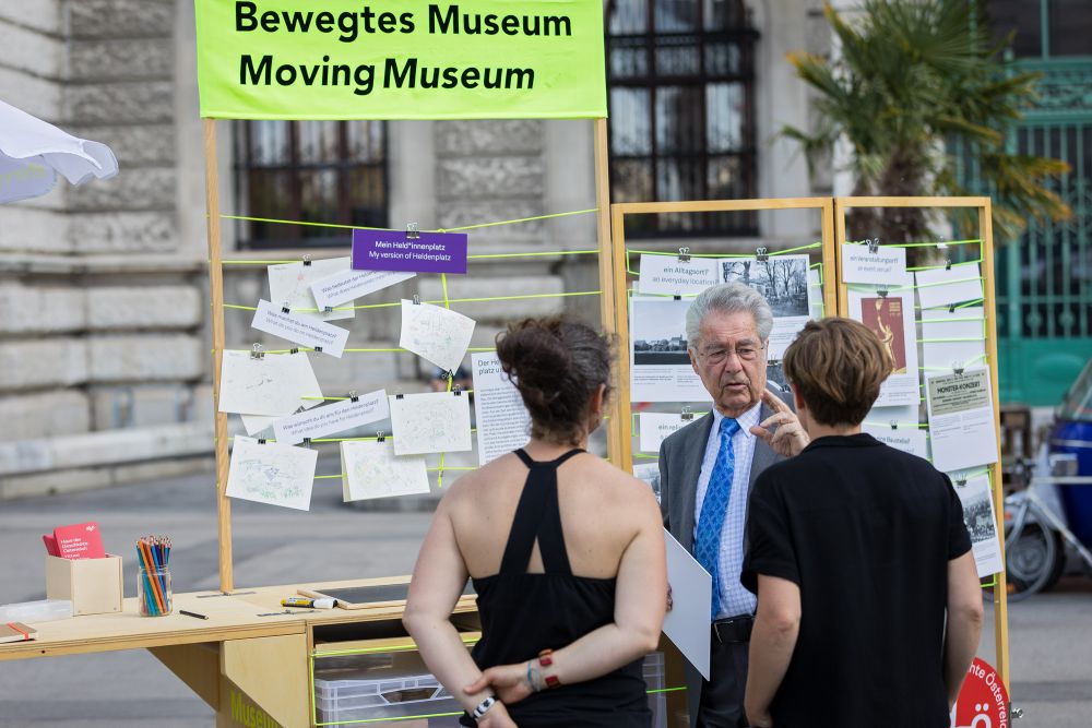 Prof. Heinz Fischer talks with two young women. They are standing in front of the wooden cart, with a banner of Moving Museum above it. The cart consists of folding wooden frames, which hold small pictures and scraps of paper. In the background, the neo-classical building of the Austrian House of History.