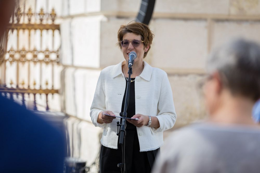 Photo taken from behind the audience. A middle-aged woman speaks to the microphone. She wears a white jacket, a black dress, and tinted glasses. White neo-classical gate in the background.