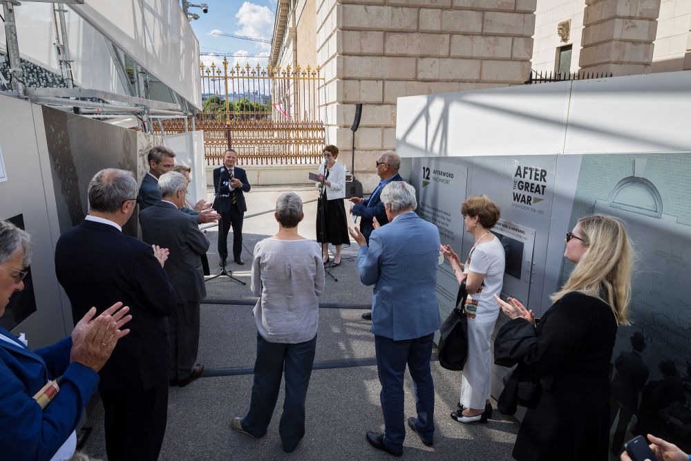 A small crowd of dignitaries gathers in a narrow space between the two elements of the After the Great War installation. In front of them, a man and a woman speak to the microphones. Neo-classical gate in the background.