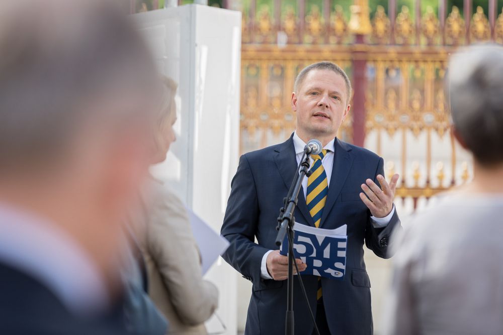 Photo taken from behind the audience. A middle-aged man speaks to the microphone. He is dressed in a suit and blue and yellow tie. holds a folder with ENRS logo.
