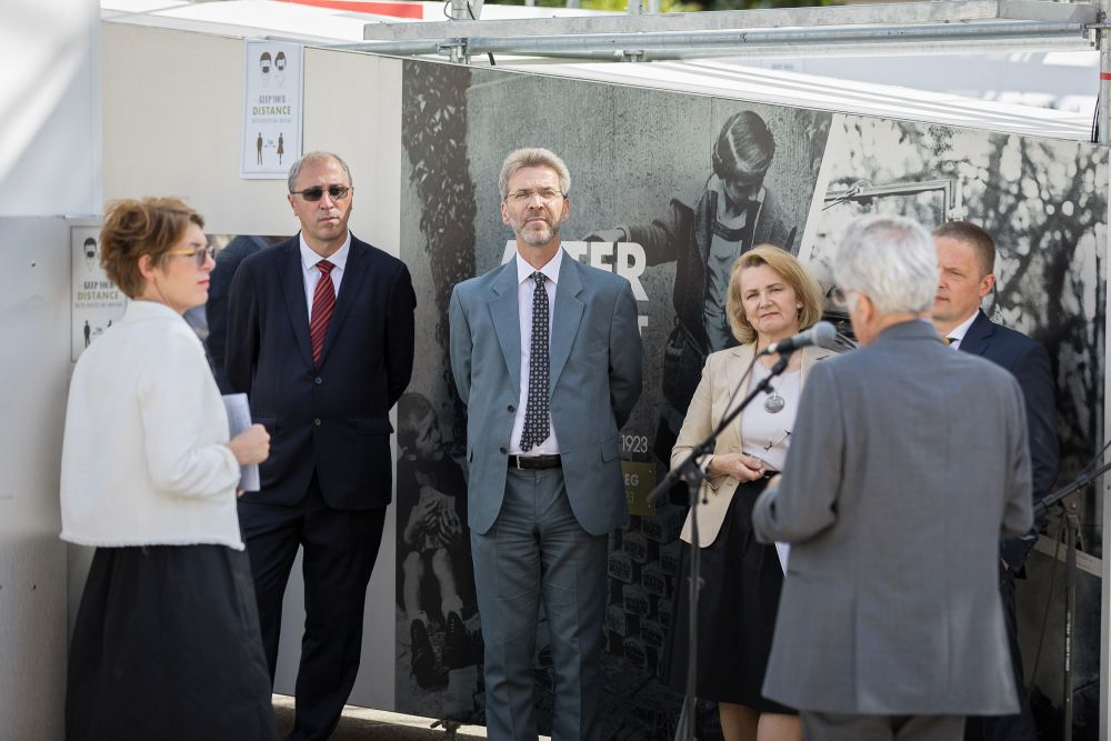 A group of five well-dressed dignitaries faces the camera, listening to the man speaking to the microphone. They are standing on a backdrop of the outer wall of the exhibition, decorated with a black-and-white photo collage.