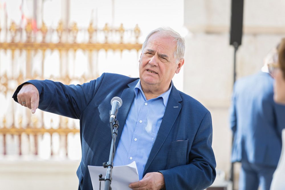 Prof. Arnold Suppan speaks into the microphone, gesturing with his hand. He is facing the camera. In background, the neo-classical white and gold fence of the Heldenplatz.