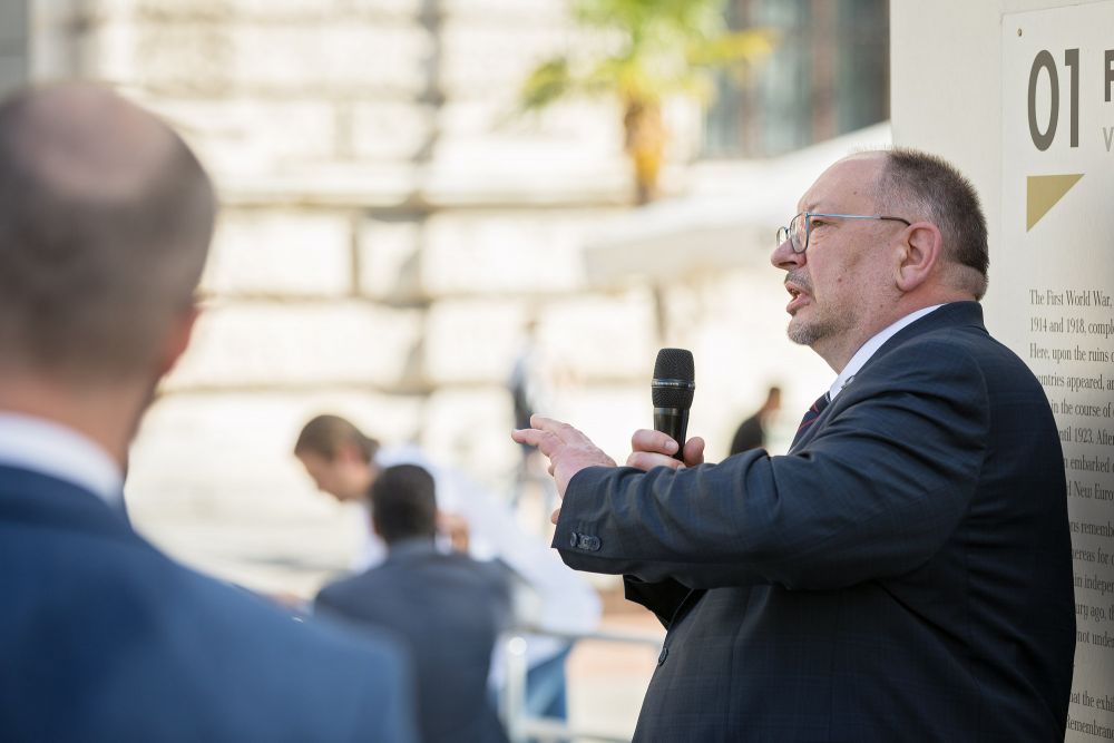 Prof. Jan Rydel holds a microphone and speaks with visible enthusiasm. His back faces one of the walls of the After the Great War exhibition. In the background, the neoclassical architecture of Hofburg and out-of-focus silhouettes of passers-by.