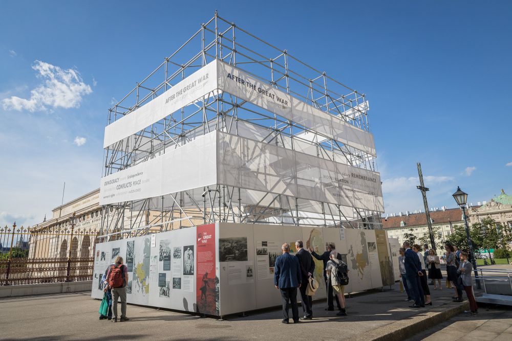 Sunny day.  Cube-like installation of the After the Great War exhibition stands in the square, groups of people gather around it, reading from infographics presented on its walls. In the background, the neo-classical architecture of the Heldenplatz.