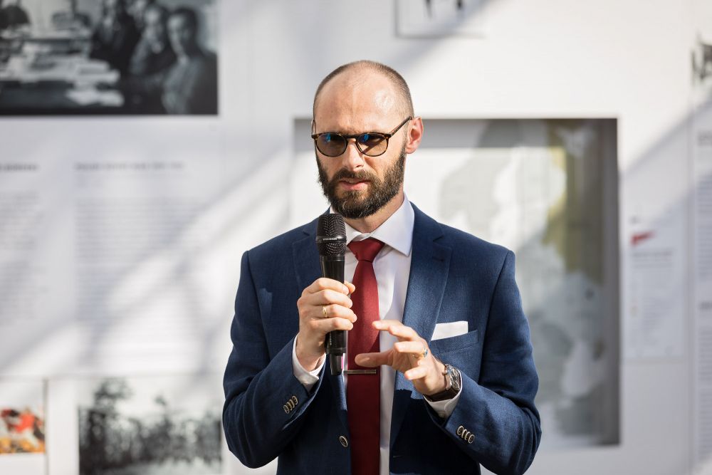 Dr. Bartosz Dziewanowski-Stefańczyk, Deputy Head of ENRS Academic Section speaks into the microphone, gesticulating with his hand. He faces the camera. Behind him, an outer wall of the After the Great War exhibition, with infographics and a map of war-torn Europe of 1918.