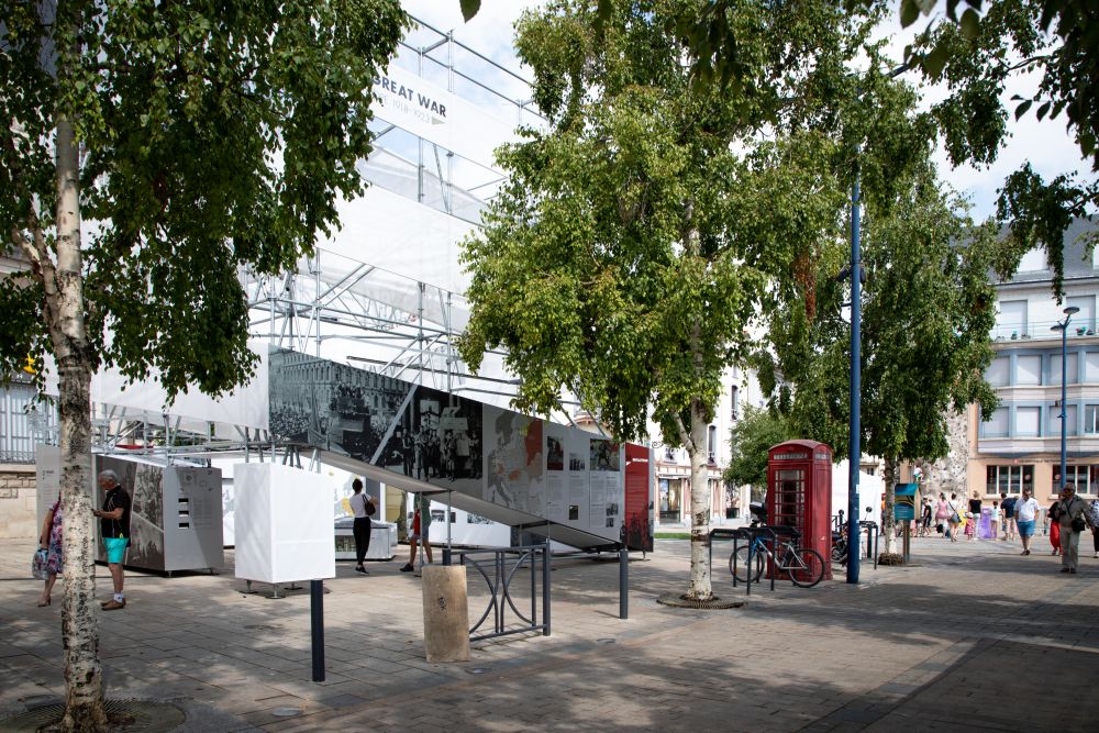 Sunny day. The cube-like installation of the After the Great War exhibition stands in the middle of a busy square. groups of people pass by or stop at the exhibition. It is surrounded by a row of birch trees.
