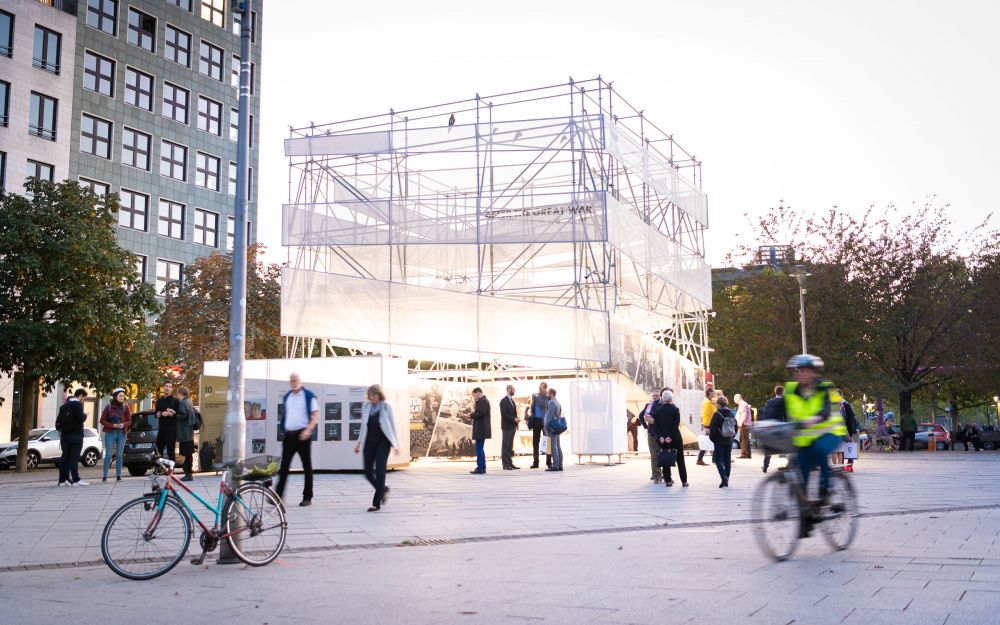 The cube-like installation of the After the Great War exhibition stands in the middle of a busy square. Around it, multiple people pass by or stop to chat or inspect the installation. A cyclist in a high-vis vest rides by the camera. Greenery in the background.