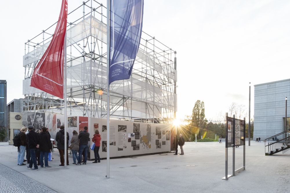 Sunny day. The cube-like installation of the After the Great War exhibition stands in the middle of a square. Two large flagpoles stand in front of it, with blue and red banners waving in a wind.  A small group of people gathered next to it.