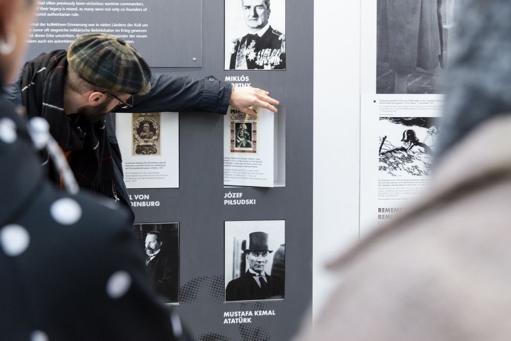 Interior of the cube-like installation of the After the Great War exhibition. A man stands in front of on of the walls, on which the portraits of the leaders of the interwar period are presented. He spins one of them, revealing the text of biographical note behind it.