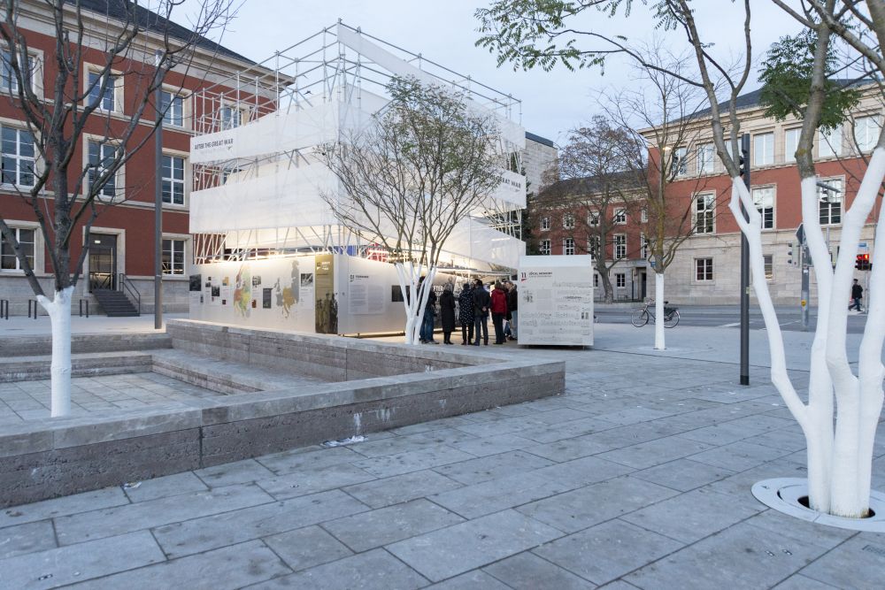 The cube-like installation of the After the Great War exhibition stands in the middle of a square. It is surrounded by small trees and red-brick buildings. A small group of people gathered next to it.