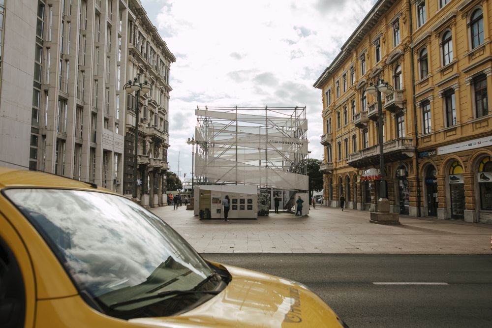 In the foreground, a yellow car. Further in the background, the cube-like installation of the After the Great War exhibition stands in the middle of an empty square.