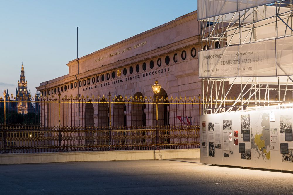 Early dawn. Illuminated cube-like installation of the After the Great War exhibition stands in the square on the left. On its right, decorative black and gold fence. Further in the background, a neoclassical gate to Heldenplatz.