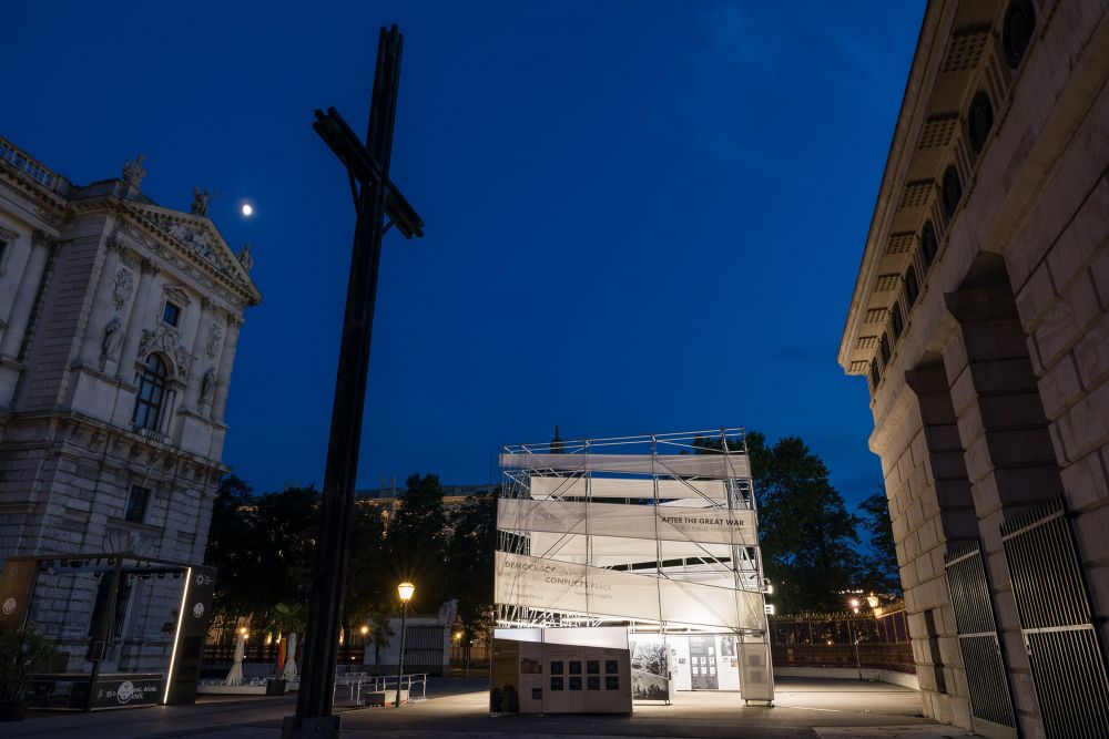 Night. Illuminated cube-like installation of the After the Great War exhibition stands in the square, framed by the neo-classical palace on the right and gate on the left. Next to it stands a large wooden cross.