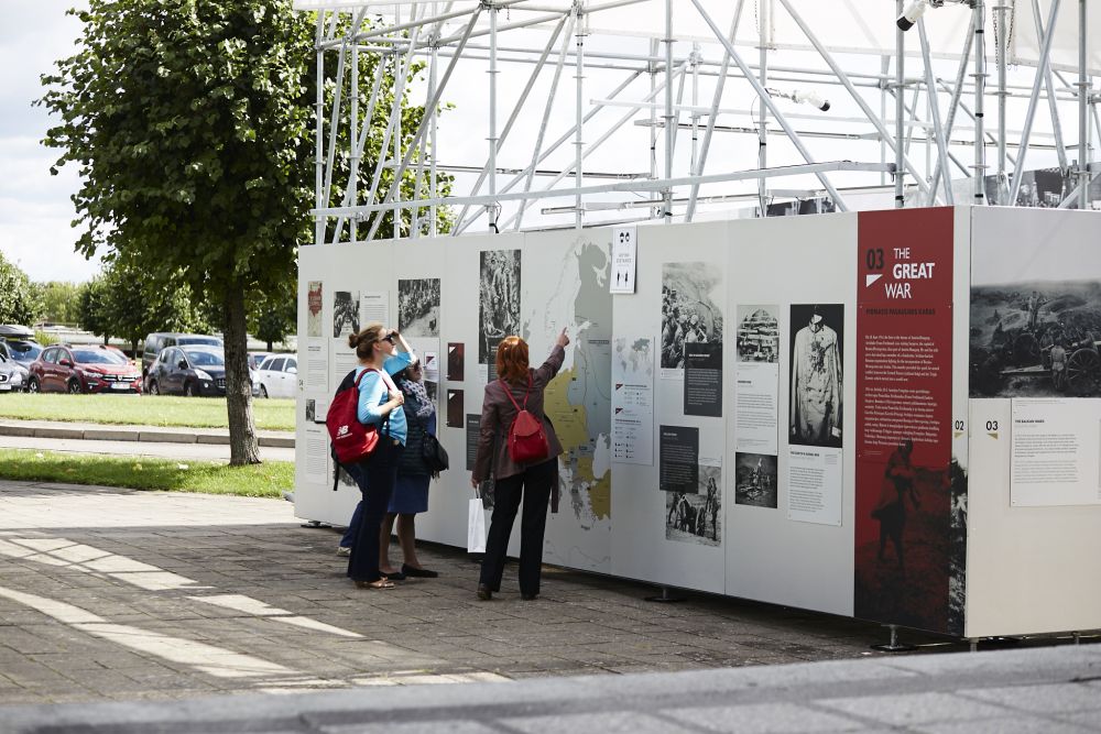 A group of women in front of the outer wall of the After the Great War installation. One of them points towards the map presented on one of the panels, depicting the extent of the eastern front of the First World War. Other women look at it with interest.