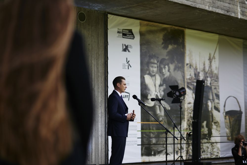 A well-dressed man stands in front of a concrete modernist building. He speaks to the microphone. A large poster advertising the exhibition hangs on a facade of the building.