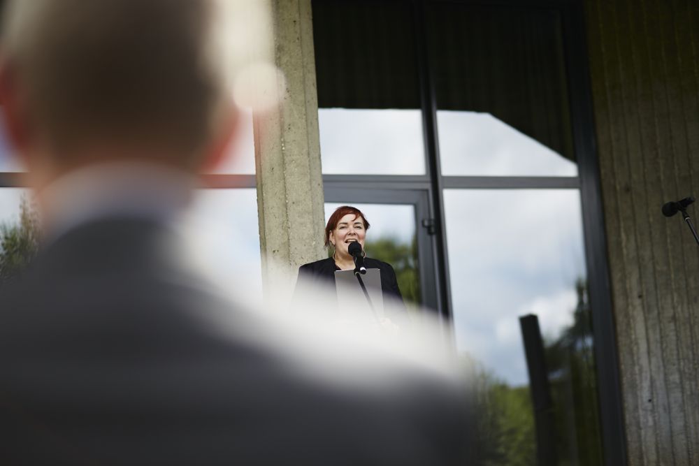 Photo taken from behind the audience. A woman  speaks to a microphone in front of a concrete modernist building.