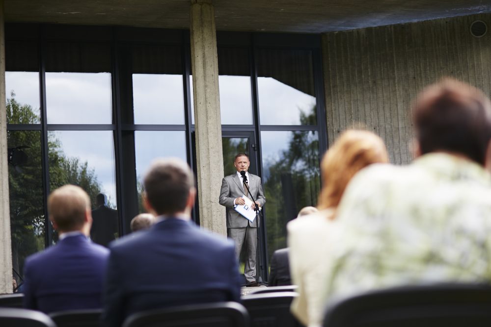 ENRS director Rafał Rogucki stands with a microphone in front of a concrete modernist building. Facing the camera, he is addressing a small crowd of dignitaries.