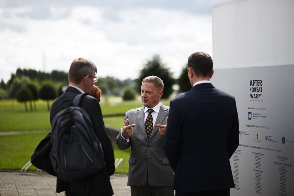 A man in a grey suit is speaking to two men and a woman gathered around him. On the right, is an outer wall of the After the Great War installation. An open green space in the background.