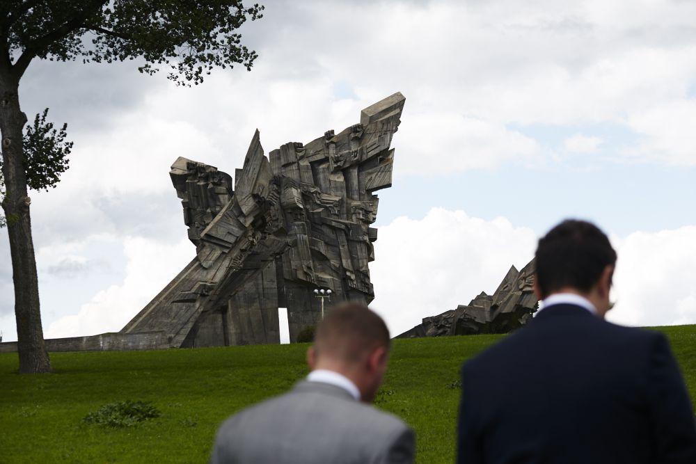 Out of focus, two men face back the camera. In the focus, a large modernist monument made of tilted concrete slabs in front of them.