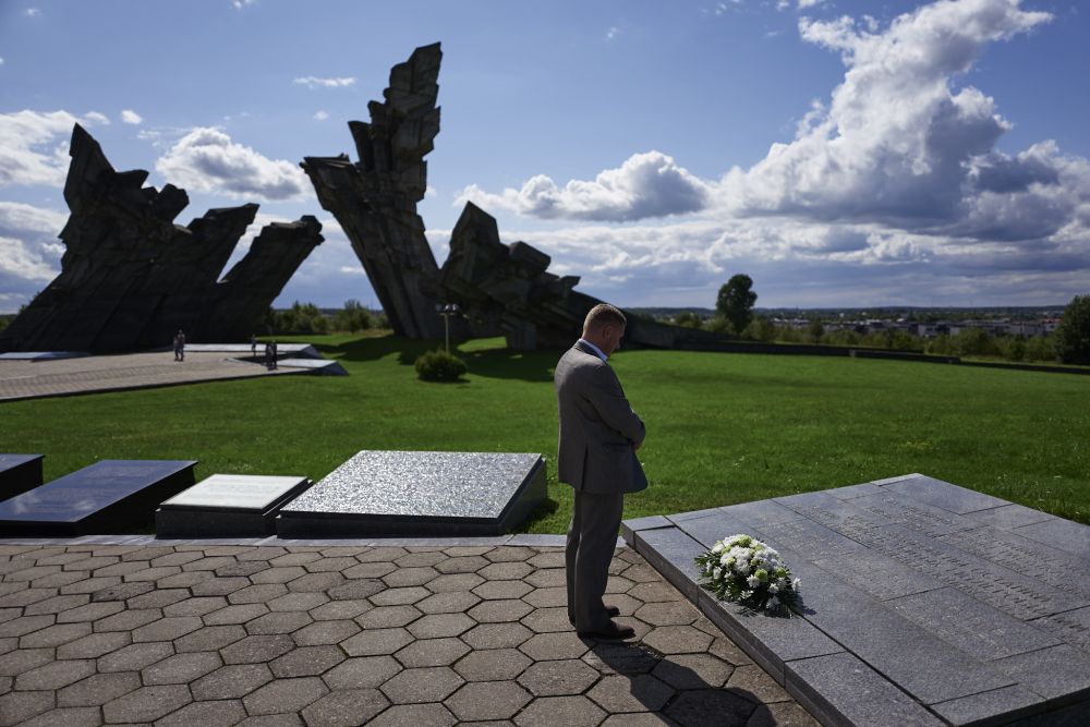 In the foreground, a single man stands solemnly in front of the commemorative plaque. A bouquet of white flowers is placed on a plaque. In the background,  a modernist monument made of concrete slabs dominates otherwise open green space.