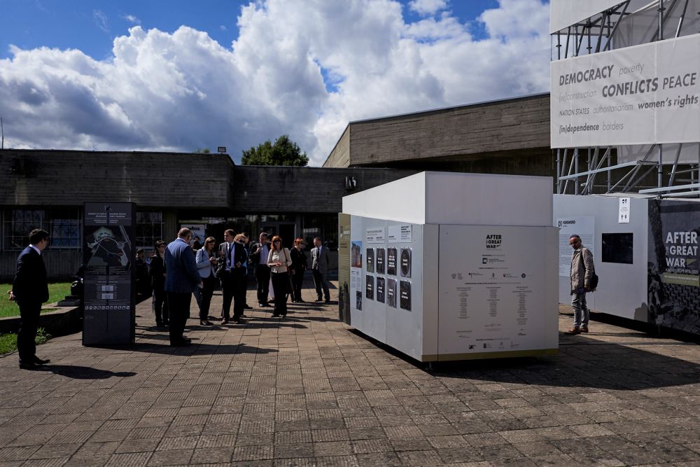 A crowd of people gathers on a small square in front of the modernist concrete building. On the right, A cube-like installation of the After the Great War exhibition.