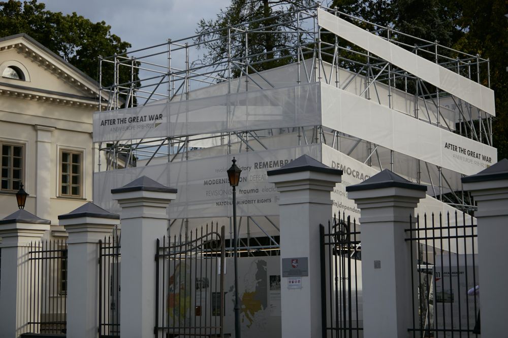 Cube-like installation of After the Great War exhibition stands behind the elegant neo-classical gate. Left to it, neo-classical white building of Tuskulėnai manor. Greenery in the background.
