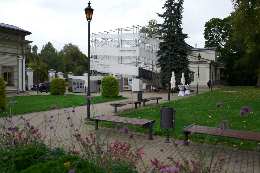 The well-kept garden with green grass, flowers, and wooden benches. In the foreground, neo-classical white buildings of the Tuskulėnai Peace Park complex and cube-like installation of the After the Great War exhibition.