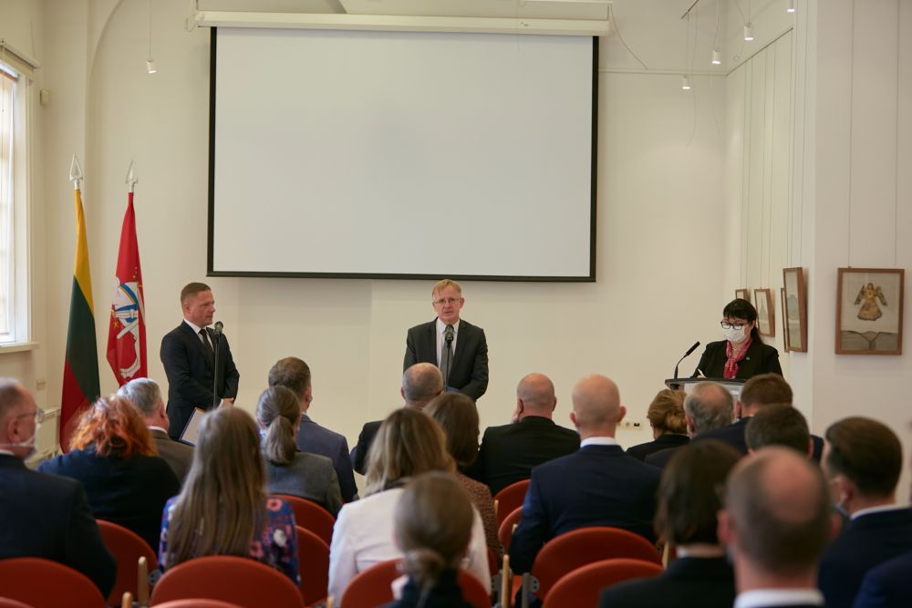 Interior, medium-sized white room. The rows of chairs densly packed with the audience. In front of them, three speakers face the camera. Men on the left and in the middle stand up, while the woman on the speaks from behind the lectern. Projector board and two Lithuanian flags in the background.