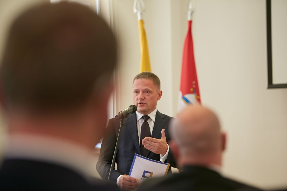 Photo taken from behind the audience. A middle-aged man speaks into the microphone. He holds a folder with ENRS logo. Projector board and two Lithuanian flags in the background.