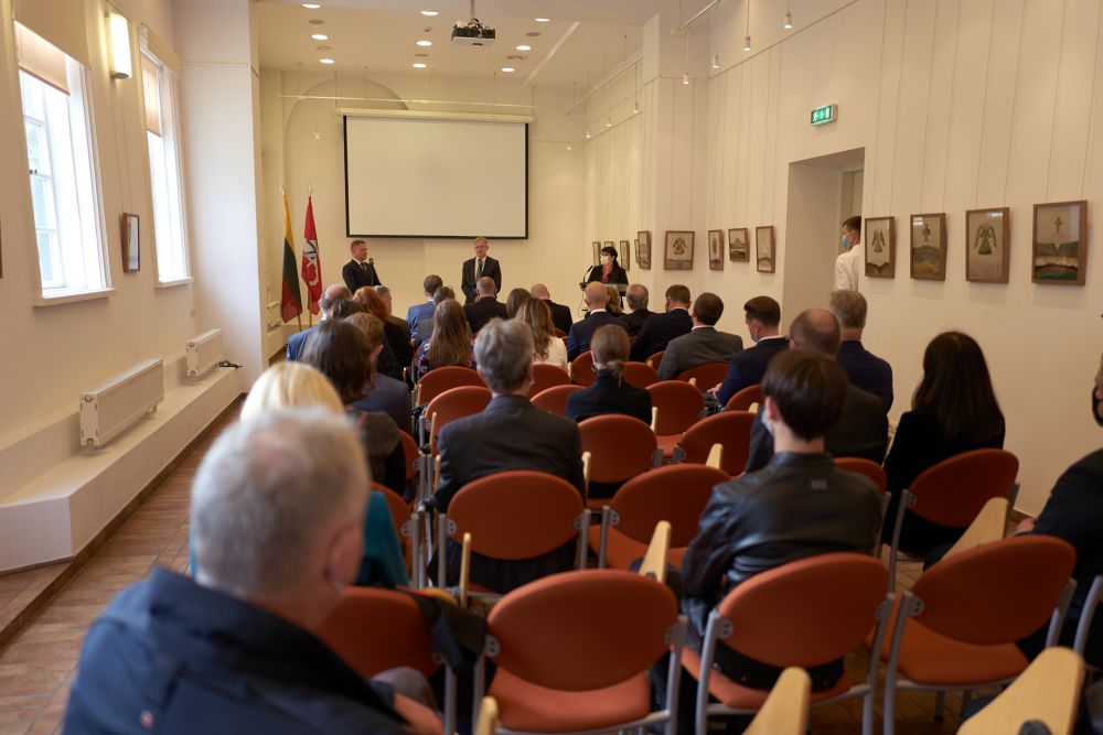 Interior, medium-sized white room. The rows of chairs densly packed with the audience. In front of them, three speakers face the camera. Men on the left and in the middle stand up, while the woman on the speaks from behind the lectern. Projector board and two Lithuanian flags in the background.