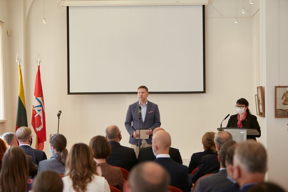 Interior, medium-sized room. The middle-aged man facing the camera speaks in front of a packed auditorium. On his left, a woman behind the lectern, possibly translating his speech. Projector board and two Lithuanian flags in the background.