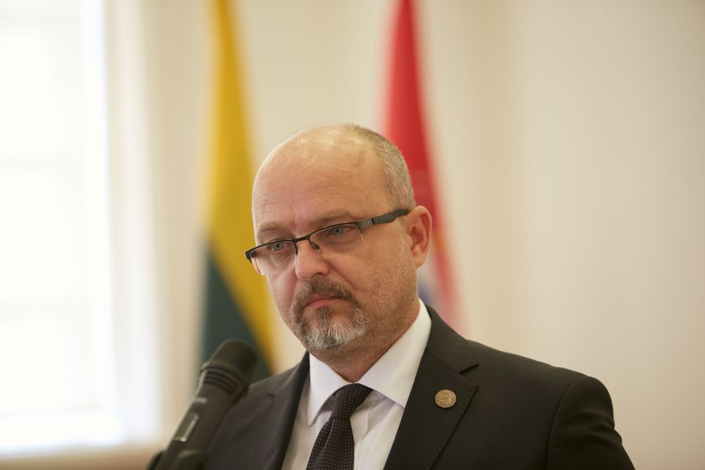 A close-up of the middle-aged man. Dressed in a suit and glasses stands in front of a microphone, facing a camera. Out of focus, two Lithuanian flags are visible in the background.