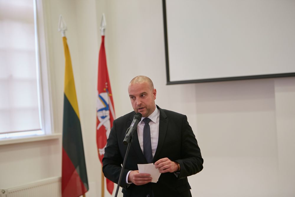 A close-up of the middle-aged man. Dressed in a dark suit he  speaks to a microphone, facing a camera. Projector board and two Lithuanian flags in the background.