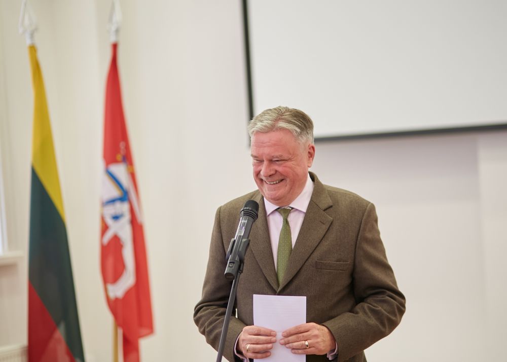 A close-up of the middle-aged man. Dressed in a brown suit he stands in front of a microphone, facing a camera and smiling. Projector board and two Lithuanian flags in the background.