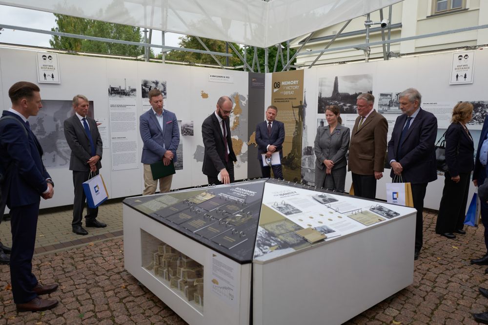 Interior of the installation of After the Great War exhibition. In the center, a knee-high board with the infographics, surrounded from all sites by the walls of the exhibition. A young man in the center points towards something on a board. Surrounding dignitaries look at it attentively.